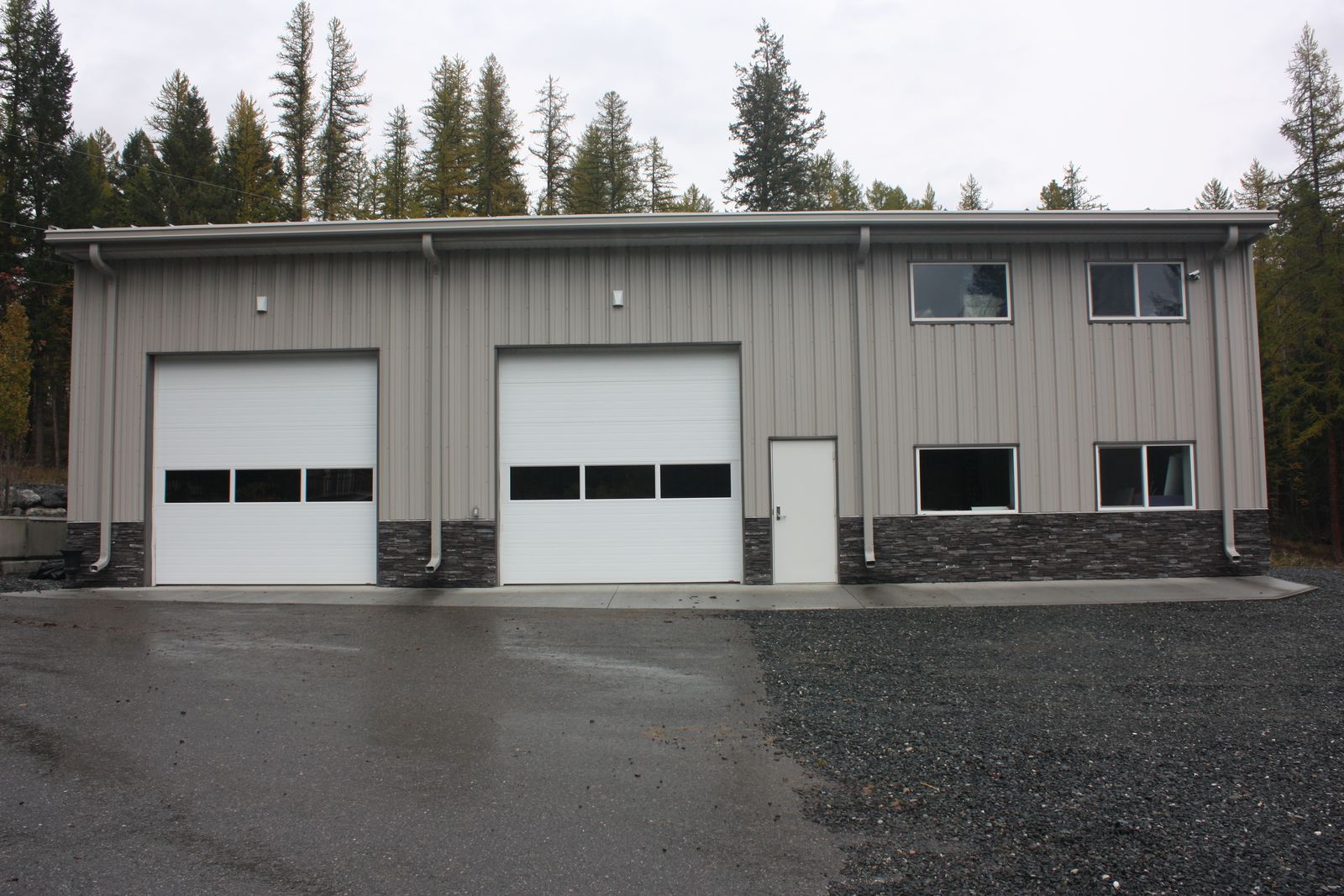 Grey steel shop with two white overhead doors, a stone base and a gravel apron