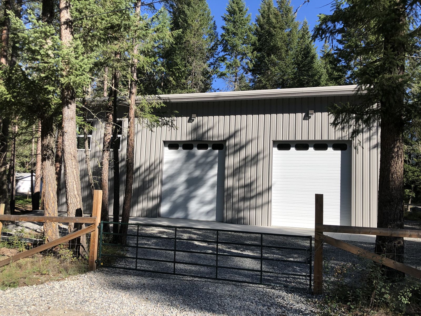 Grey steel shop with two overhead doors behind a wooden fence and gate, with evergreen trees