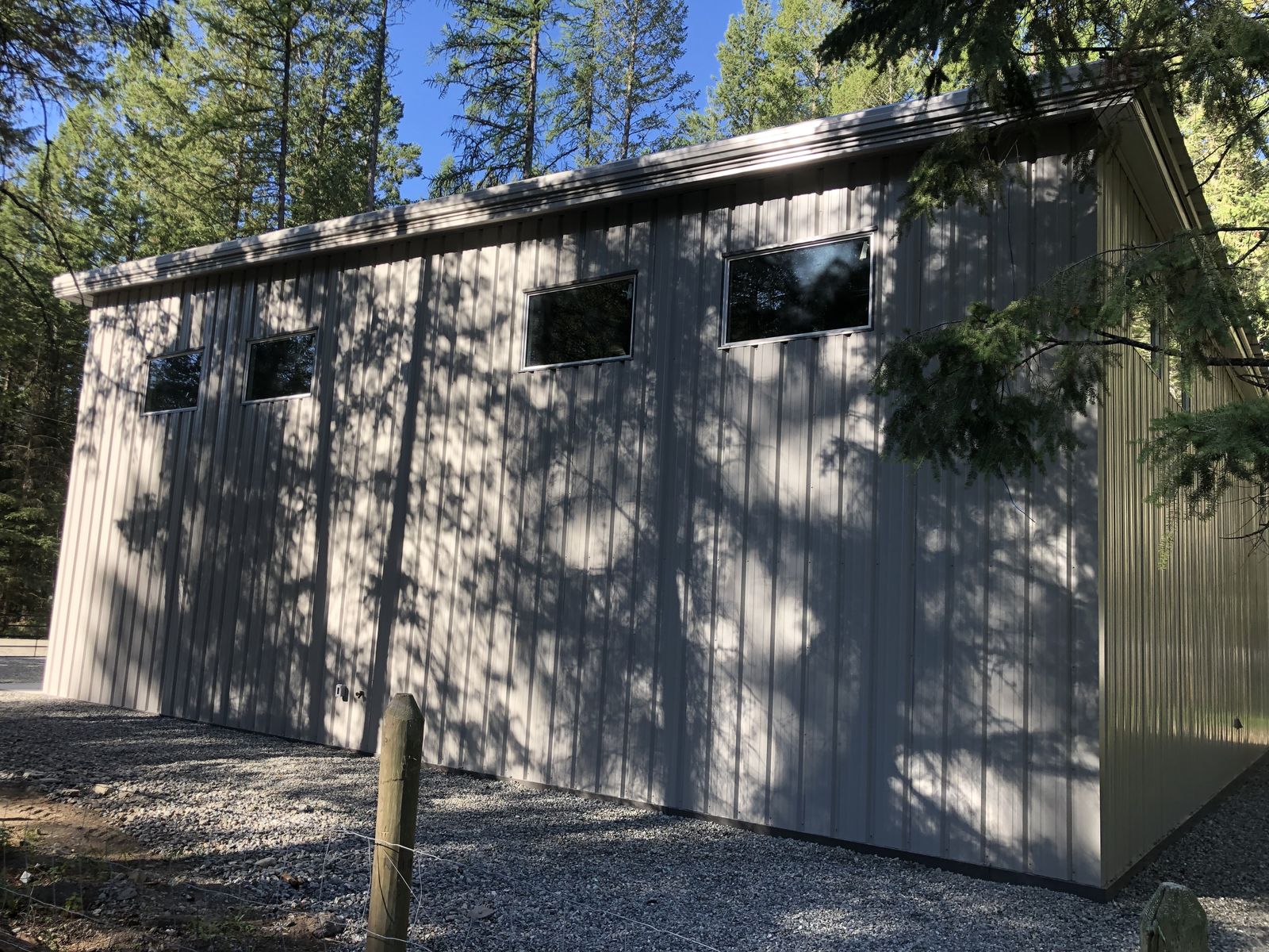 Side wall of a tall grey steel shop with windows, surrounded by trees